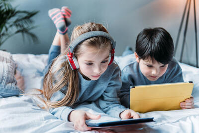 little girl in headphones and boy using digital tablets while lying on bed