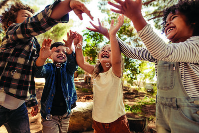 Group of kids playing with soap bubbles in forest. Boy blowing soap bubbles with friends trying to catch the bubbles.