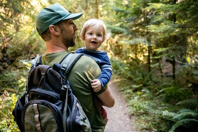 A dad carries his one year old son while enjoying a sunny hike in the Pacific Northwest.  Evergreen trees and ferns line the trail on the Washington state peninsula.
